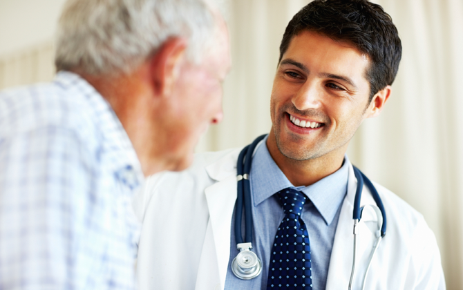 A male physician converses with an elderly male while providing home nursing care.