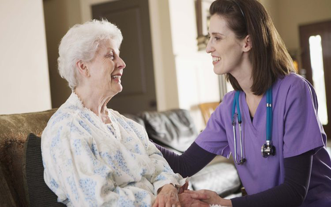 An older woman is being cared & supported by a nurse who is talking to her attentively.