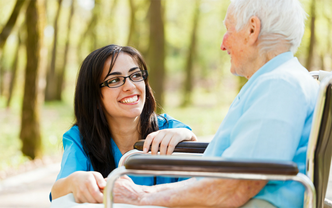 A personal home care assistant converses with a senior citizen in a wheelchair with her smiley face.