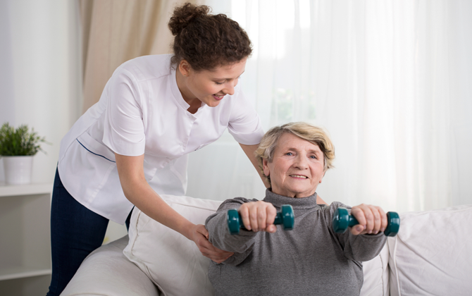 A respite home care helps exercise with dumbbells while attentively supporting an elderly woman