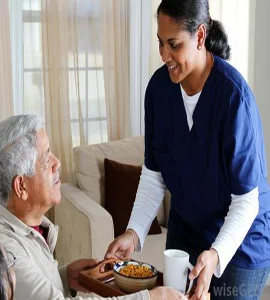 An elderly guy is given lunch by a nice nurse, who makes sure he eats and stays well